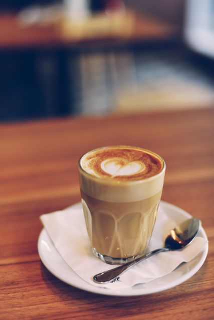A close-up view of a latte in a glass cup placed on a white napkin and saucer, with a spoon beside it on a wooden table. The latte features heart-shaped foam art, indicating a relaxed and cozy coffee shop setting. Ideal for use in cafe menus, coffee shop advertisements, lifestyle blogs on morning routines, and marketing materials for hot beverages.