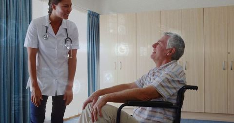 Nurse leaning and talking with elderly man in wheelchair, caring conversation in ward