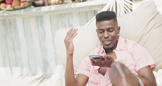 Young man lounging on hammock while using smartphone in outdoor setting. Suitable for concepts related to leisure, technology, communication, summer relaxation, remote work, and casual lifestyle.