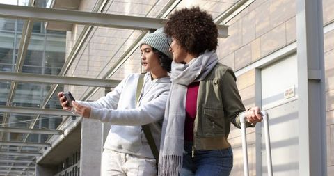 Diverse women checking phone at airport concourse while pulling suitcase under glass canopy
