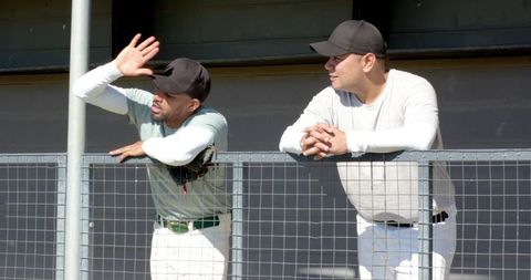Two baseball teammates leaning on dugout railing shielding eyes while watching sunny outdoor game