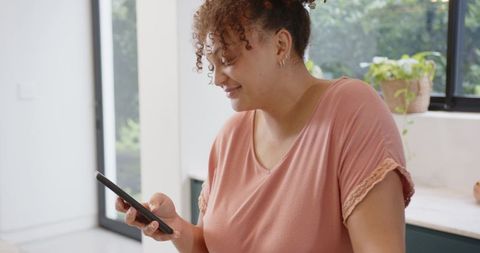 African american woman leaning on kitchen counter scrolling smartphone in sunlit kitchen