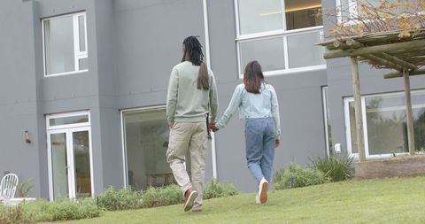 Interracial Couple Walking Hand-in-Hand Toward Modern Gray Home With Pergola and Lawn