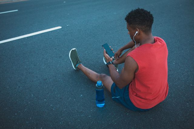 African American man sitting on a coastal road, using a smartphone and wearing headphones. He is dressed in sportswear, indicating he is taking a break from a workout or run. A water bottle is placed beside him, emphasizing the importance of hydration during exercise. This image can be used for promoting fitness, healthy lifestyles, outdoor activities, and sports-related content.