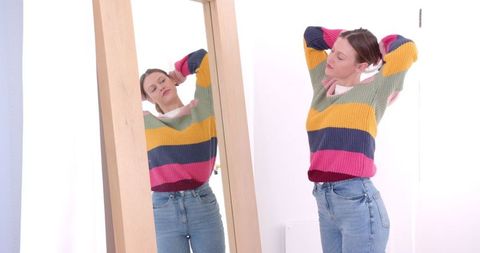 Woman adjusting hair in colorful sweater by large mirror at home