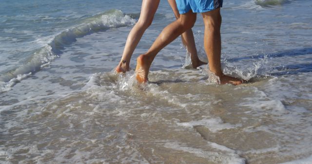 Two people are enjoying a walk along the shoreline, with their feet splashing in the shallow ocean water, with copy space. Capturing the simplicity of a beach stroll, this image evokes the carefree moments of summer leisure.