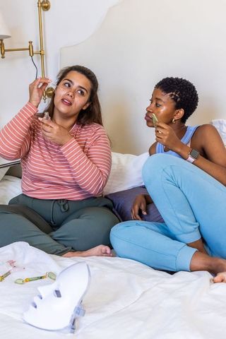 Two friends applying facial serum and jade roller on bed, cozy self-care skincare routine