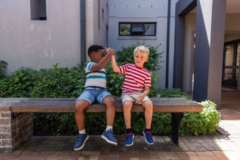 Diverse boys high-fiving on wooden bench in sunny brick courtyard, playful summer friendship