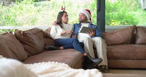 Diverse couple relaxing on sofa wearing Santa hat and reindeer antlers using tablet