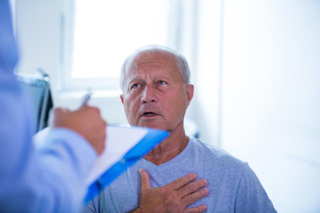 Senior man consulting with a doctor in a hospital setting. The man appears to be discussing his symptoms, with a hand on his chest, while the doctor takes notes. This image can be used for healthcare, medical consultation, patient care, and elderly health-related content.