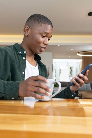 African american man checking phone while holding coffee in modern kitchen