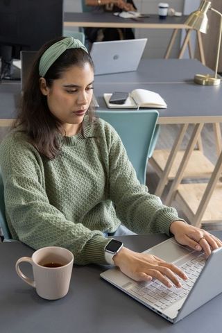 Asian woman working on laptop in modern coworking space with coffee cup and smartwatch