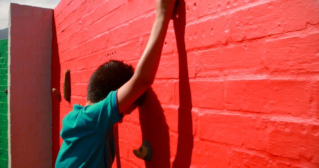 A young African American child is engaged in outdoor rock climbing on a bright red wall, with copy space. Rock climbing activities are excellent for developing strength and coordination in children.