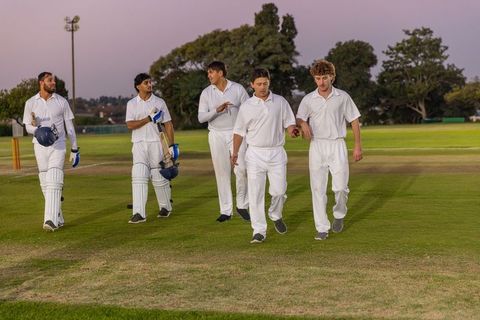 Cricket teammates walking twilight pitch carrying bats helmets and ball, teamwork outfield