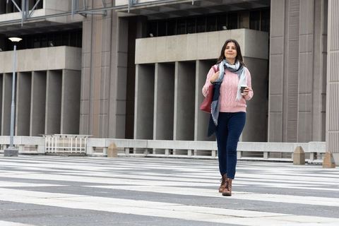Middle-aged woman walking confidently across urban plaza crosswalk holding coffee