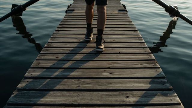 Walking man on wooden dock with oars and long shadows over calm reflective water at sunset