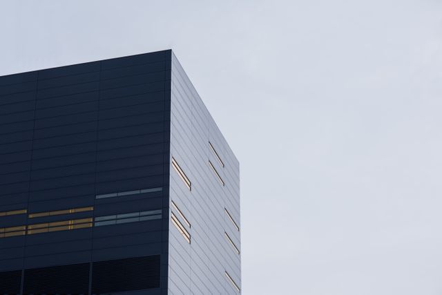 Image shows a portion of a modern skyscraper with sleek, glass architecture. The building is part of a cityscape and is set against a cloudy sky. Ideal for business promotions, corporate presentations, urban development context, and architectural portfolios.