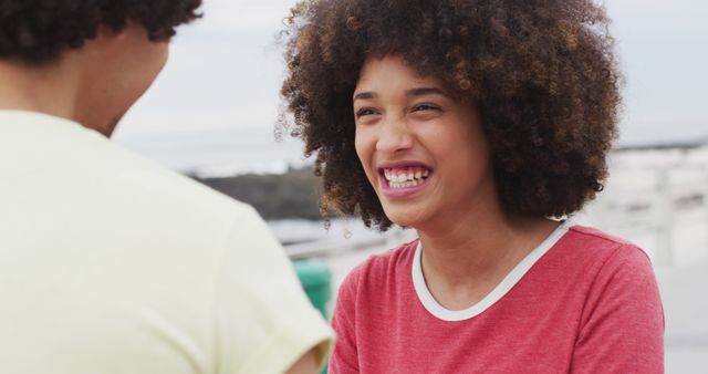 A young woman with curly afro hair laughs while engaging in conversation with a friend at the beach. Both are casually dressed, creating a relaxed and joyful atmosphere. Ideal for use in advertisements, social media posts, and websites promoting friendship, outdoor activities, and happiness.