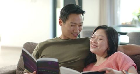 Asian couple reading together on sofa at home, sharing cozy relaxed afternoon moment