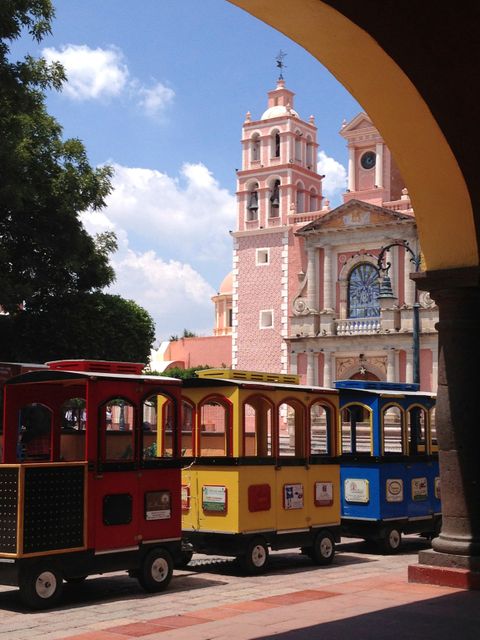 Bright tourist train parked in front of a beautiful, historic church on a sunny day. Blue sky with clouds adds to picturesque scene. Ideal for topics related to tourism, cultural exploration, city trips, and heritage sites.