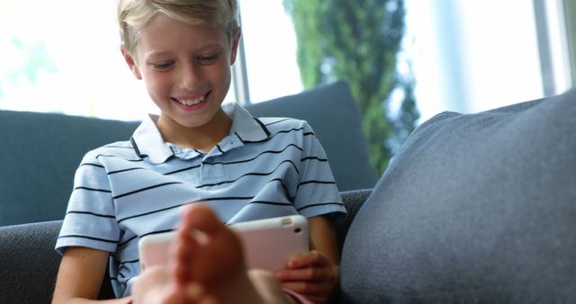 Young boy sitting on couch using tablet device, smiling happily. Casual home environment, ideal for promoting technology use among children, online learning, or family life. Exhibits positive emotions and comfort in a domestic setting.