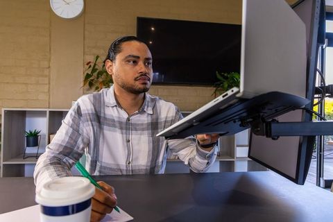 Young man adjusting laptop on monitor arm while taking notes in daylight workspace