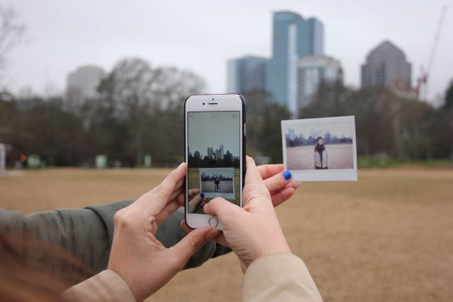 Hands holding smartphone and an instant photo against urban park background with city skyline. Usage ideas include photography concepts, city lifestyle, social media, and capturing moments in nature.