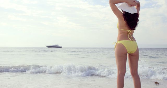 Woman wearing yellow bikini and sun hat enjoying the beach with a boat on the horizon. Great for travel and tourism promotions, summer and beach themes, and advertising swimwear.