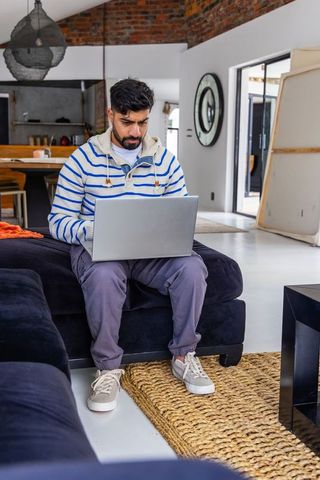 Indian man working on laptop in modern loft living room with exposed brick and cozy sofa