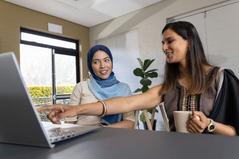 Two professional women collaborating at laptop in modern home office, hijab and embroidered tunic