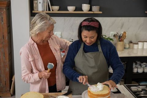 Mature women frosting layer cake together in cozy home kitchen, baking friendship