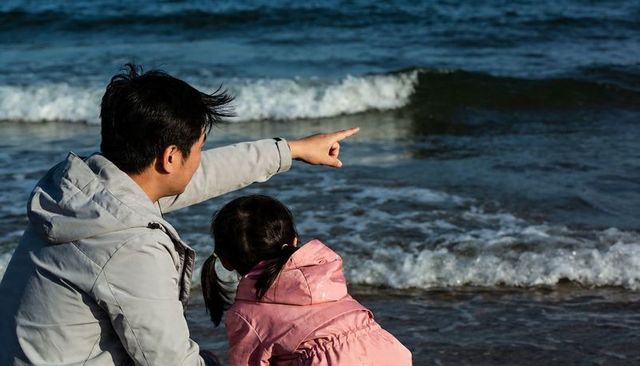 Father and daughter watching ocean waves on sandy shore, father pointing toward horizon