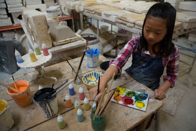 Young girl painting a ceramic bowl in a pottery workshop. She is surrounded by various paints, brushes, and pottery tools. Ideal for use in educational materials, art and craft promotions, children's activities, and creative hobby advertisements.