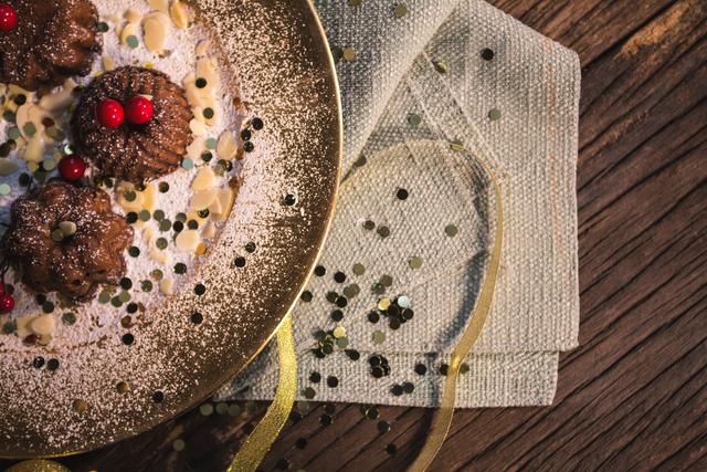 Mini chocolate bundt cakes dusted with icing sugar on a gold plate. Red berries add a festive touch. Ideal for holiday celebrations, party invitations, food blogs, and dessert menus. Rustic wooden table and gold confetti enhance the festive atmosphere.