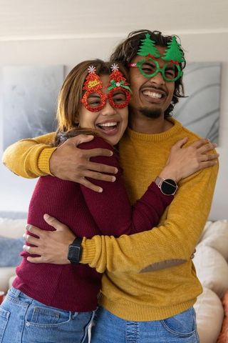 Young couple hugging and laughing wearing festive glasses and cozy knit sweaters