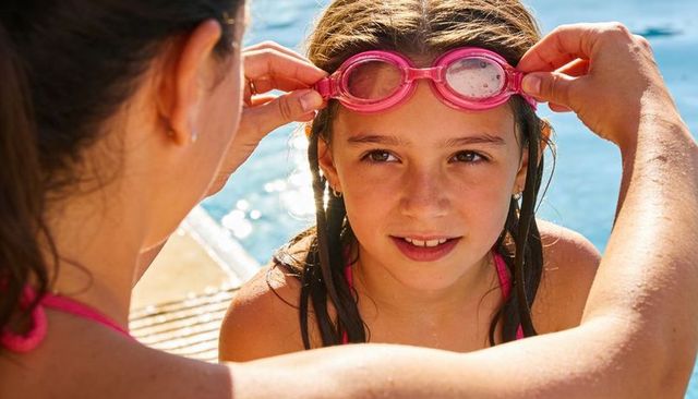 Caregiver adjusting pink swim goggles on young girl at sunny poolside with blue water