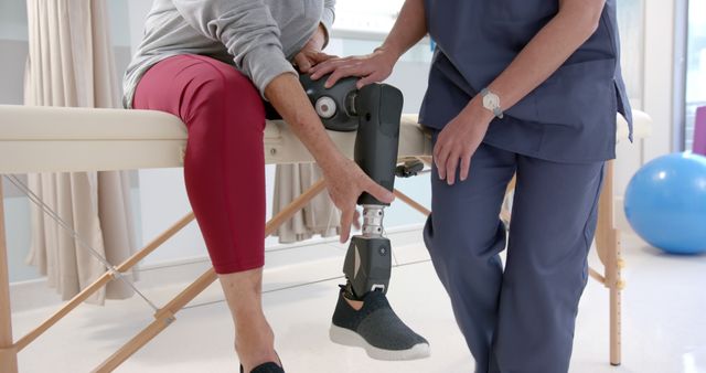 A physical therapist in blue scrubs adjusting prosthetic leg for a disabled woman in a clinic. Woman wearing maroon pants and black shoe. Image can be useful for articles, blog posts, and health websites focusing on medical interventions, prosthetics technology, rehabilitation services, and healthcare professional assistance.