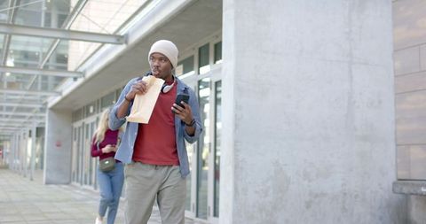 African american man walking, eating from paper bag and checking phone under glass canopy