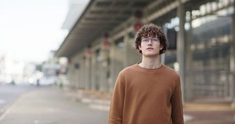 Young man with curly hair and glasses standing at modern transit terminal in brown sweater
