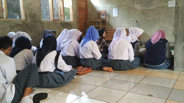 Group of high school students wearing uniforms sitting on the floor in a classroom, engaging in a group discussion. The spacious room has a rustic look with concrete walls and tiled floor. Ideal for educational content, articles about teamwork in schools, or discussions on traditional teaching environments.