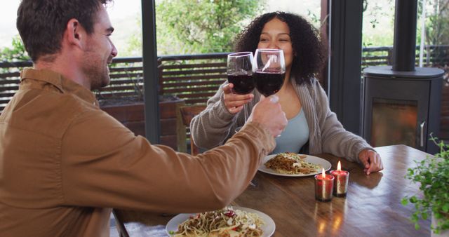 A couple is having a romantic dinner, toasting with wine glasses while sitting at a wooden table with plates of pasta and lit candles. This image suits content focused on relationships, dining, lifestyle articles, date night ideas, and hospitality services promoting a cozy dining experience.