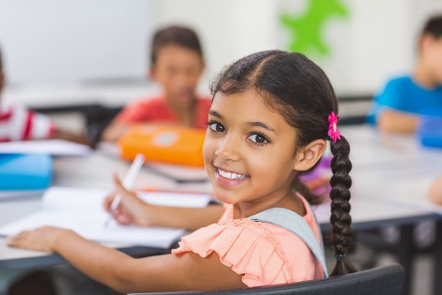 Young girl smiling while writing in classroom, ideal for educational brochures, websites, and teacher blogs promoting a joyful learning environment. Perfect for illustrating concepts of education, student engagement, and diversity in schools.