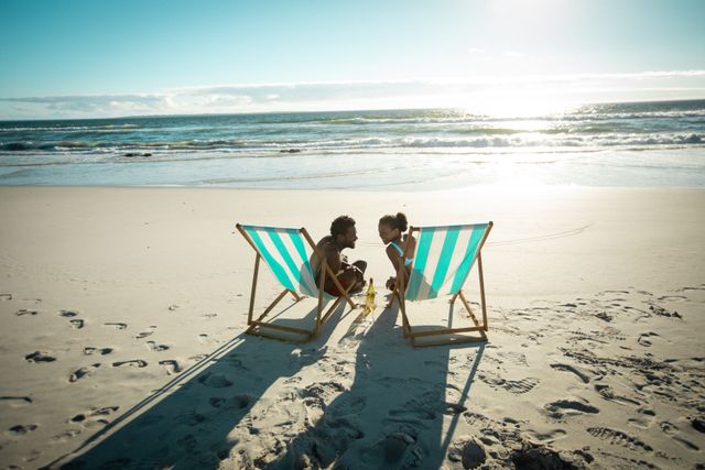 Happy african american couple sitting in deckchairs talking with drinks on sunny beach by the sea. holiday, romance and leisure time at the beach.
