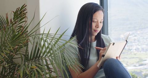 Asian woman writing on tablet at window with city view, relaxed home workspace and palm plant