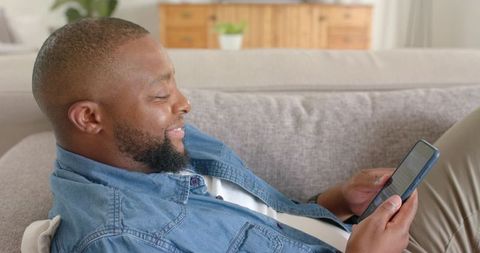African american man relaxing on light-gray sofa scrolling smartphone and smiling at home