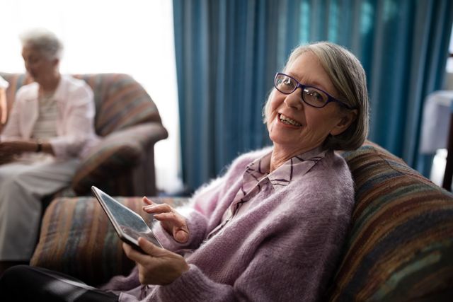 Portrait of smiling senior woman sitting with digital tablet on armchair at retirement home