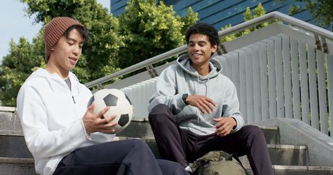 Two young men sitting on outdoor steps holding soccer ball and laughing, campus sports