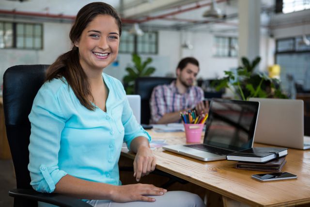 Business woman smiling while working at her desk in a modern office environment. Ideal for use in corporate websites, business presentations, and promotional materials highlighting professional workspaces and teamwork.