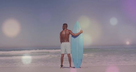 Surfer Standing with Blue Board on Sandy Beach with Ocean Waves