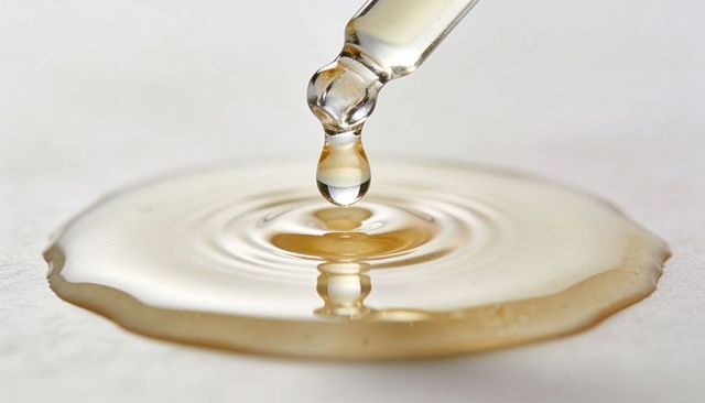 Amber droplet falling from glass dropper creating concentric ripples on glossy surface